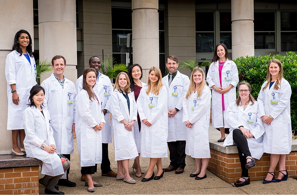 Outdoor group photo of 13 smiling women and men wearing white lab coats.